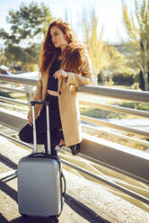 Nachdenkliche Geschäftsfrau steht mit ihrem Gepäck auf einem erhöhten Gehweg am Flughafen während eines sonnigen Tages - EHF00413