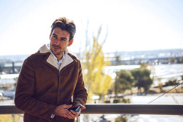 Thoughtful businessman holding smart phone while standing on elevated walkway during sunny day - EHF00415