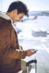 Happy businessman using smart phone by window at airport departure area - EHF00448