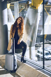 Confident businesswoman standing with luggage by window at airport departure area - EHF00454