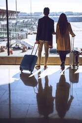 Business couple holding luggage while looking through window at airport departure area - EHF00457