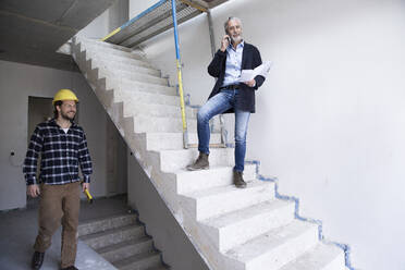 Männlicher Architekt, der über sein Smartphone spricht, während ein Bauarbeiter in einem im Bau befindlichen Haus arbeitet - MJFKF00423