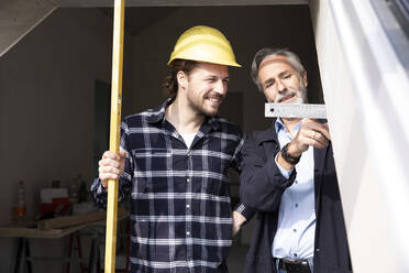 Lächelnder Bauarbeiter, der die vom Architekten beim Bau eines Hauses gezeigten Maße durch ein Fenster betrachtet - MJFKF00430