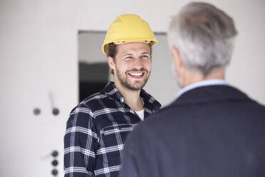 Smiling construction worker wearing hardhat talking with architect in constructing home - MJFKF00433