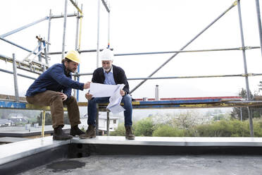 Architect and construction worker discussing blueprint while sitting on scaffold against clear sky - MJFKF00441