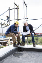 Architect and construction worker discussing blueprint while sitting on scaffold against clear sky - MJFKF00442