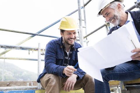 Ein lächelnder Architekt und ein Bauarbeiter besprechen einen Bauplan, während sie auf einer Baustelle gegen den klaren Himmel sitzen, lizenzfreies Stockfoto