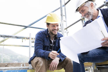 Ein lächelnder Architekt und ein Bauarbeiter besprechen einen Bauplan, während sie auf einer Baustelle gegen den klaren Himmel sitzen - MJFKF00443