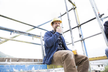 Bauarbeiter isst Essen, während er auf der Baustelle gegen den klaren Himmel sitzt - MJFKF00445