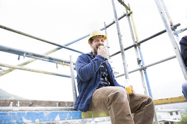 Construction worker eating food while sitting against clear sky at construction site - MJFKF00445