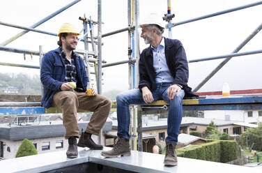 Architect and construction worker talking while sitting at construction site - MJFKF00446