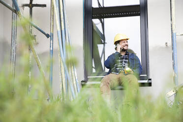 Bauarbeiter, der über sein Smartphone spricht, während er auf der Baustelle am Fenster sitzt - MJFKF00448