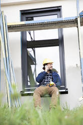 Bauarbeiter, der am Fenster sitzend über sein Mobiltelefon spricht - MJFKF00449