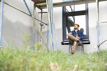 Thoughtful construction worker holding fruit while sitting against window at construction site - MJFKF00451