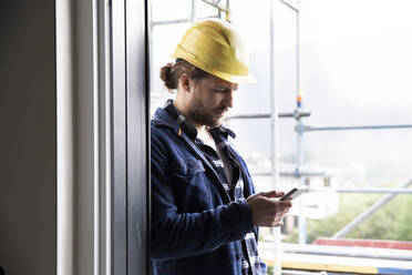 Bauarbeiter mit Helm und Smartphone am Fenster beim Bau eines Hauses - MJFKF00453