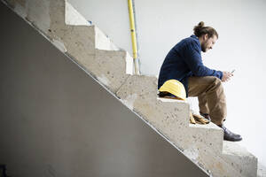 Bauarbeiter, der sein Smartphone benutzt, während er auf der Treppe eines Hauses auf der Baustelle sitzt - MJFKF00461