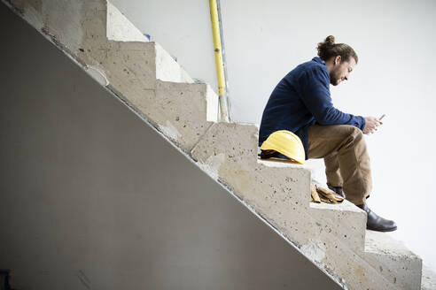 Construction worker using smart phone while sitting on steps in house at site - MJFKF00461