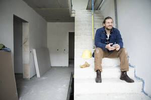 Nachdenklicher Bauarbeiter hält sein Smartphone in der Hand, während er auf den Stufen eines im Bau befindlichen Hauses sitzt - MJFKF00463