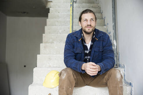 Nachdenklicher Bauarbeiter sitzt auf den Stufen eines im Bau befindlichen Hauses - MJFKF00464