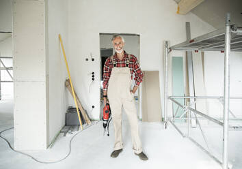 Smiling construction worker wearing overalls standing in constructing house - MJFKF00471