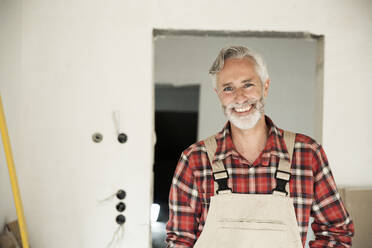 Smiling construction worker wearing overalls standing against wall in constructing house - MJFKF00472