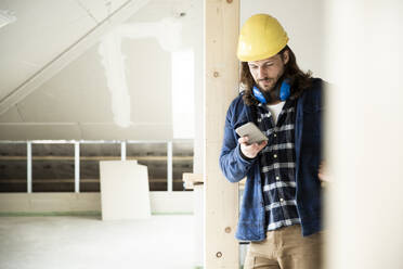 Bauarbeiter mit Helm, der sein Smartphone benutzt, während er bei der Renovierung eines Hauses am Holz steht - MJFKF00473