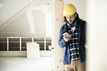 Construction worker wearing helmet using smart phone while standing by wood in renovating house - MJFKF00473