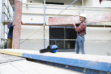 Gehörschutz auf Holz mit einem männlichen Arbeiter, der im Hintergrund Holz trägt, auf einer Baustelle - MJFKF00480