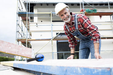 Lächelnder männlicher Architekt, der auf einer Baustelle stehend Holz trägt - MJFKF00481