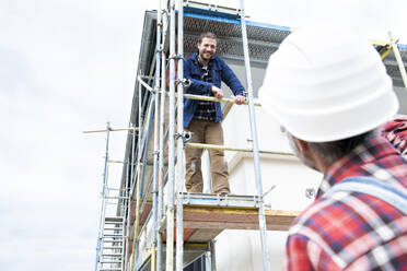 Construction worker talking with coworker while standing on scaffold at site - MJFKF00483