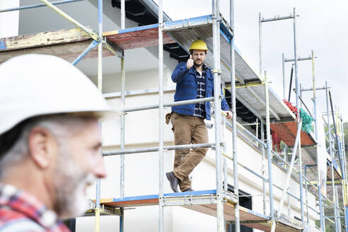 Bauarbeiter zeigt einem Kollegen den Daumen nach oben, während er auf einem Gerüst auf der Baustelle sitzt - MJFKF00485