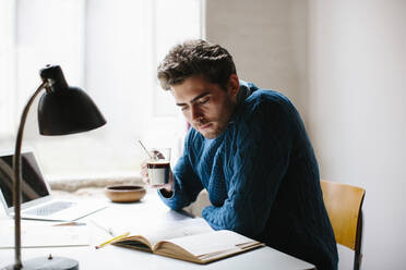 Männlicher Architekt hält Kaffee und liest ein Buch, während er am Schreibtisch im Büro sitzt - SODF00818