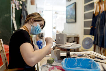 Junge Frau mit Maske bei der Herstellung von Keramik auf einer Werkbank in einer Werkstatt - EGAF00389