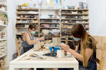 Frauen mit Masken bei der Keramikherstellung auf einer Bank in einer Werkstatt - EGAF00390