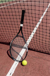 Tennis racket with ball by net in court during sunny day - EGAF00408