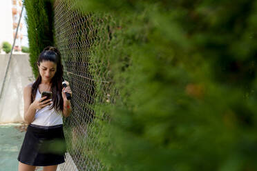 Frau benutzt Smartphone, während sie am Zaun eines Sportplatzes steht - EGAF00422