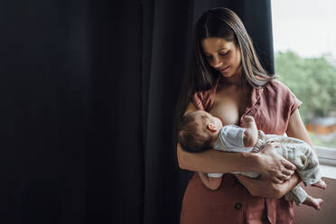 Mother breastfeeding baby boy while standing by window at home - OCMF01438