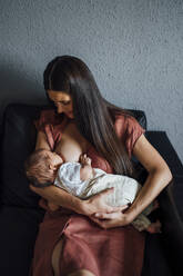 Mother breastfeeding baby boy while sitting on sofa at home - OCMF01439