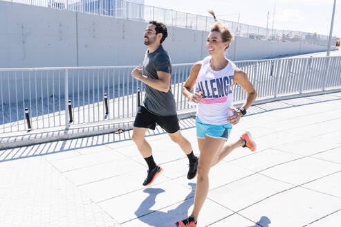 Happy fit couple jogging on city street during sunny day - JCMF00936