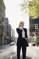 Smiling female professional holding face mask while talking on smart phone in city during COVID-19 pandemic - JOSEF00951