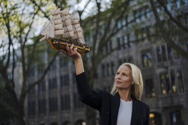 Selbstbewusste blonde Geschäftsfrau, die ein Segelbootmodell betrachtet, während sie in der Stadt steht - JOSEF00955