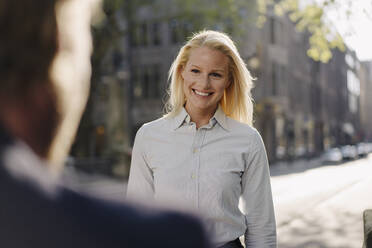 Lächelnde blonde Geschäftsfrau, die einen männlichen Kollegen ansieht, während sie in der Stadt steht - JOSEF00958