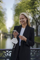 Smiling blond businesswoman holding protective face mask while using smart phone against canal in city - JOSEF00966