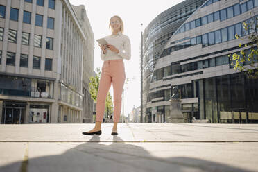 Lächelnde weibliche Fachkraft, die ein digitales Tablet hält, während sie auf einem Fußweg in der Stadt an einem sonnigen Tag steht - JOSEF00969