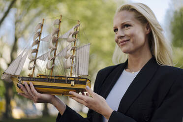 Lächelnde schöne Geschäftsfrau schaut weg, während sie ein Modellsegelboot in der Stadt hält - JOSEF00974