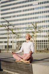 Female professional meditating while sitting in lotus position on bench against building during sunny day - JOSEF00975