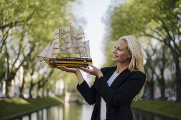 Lächelnde blonde Geschäftsfrau, die ein Modell-Segelboot betrachtet, während sie am Kanal in der Stadt steht - JOSEF00978