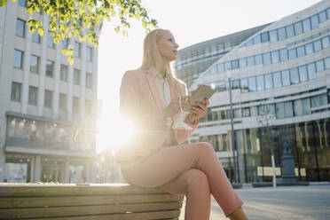 Von hinten beleuchtete Geschäftsfrau, die ein digitales Tablet hält, während sie auf einer Bank sitzt und in die Stadt blickt - JOSEF00980