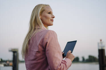 Nachdenkliche blonde Geschäftsfrau, die ein digitales Tablet hält und wegschaut - JOSEF00982