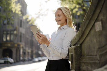 Smiling blond female professional using digital tablet while leaning on surrounding wall in city - JOSEF00983